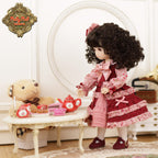 Doll in a red and pink outfit standing next to a tea set on a table with a teddy bear.