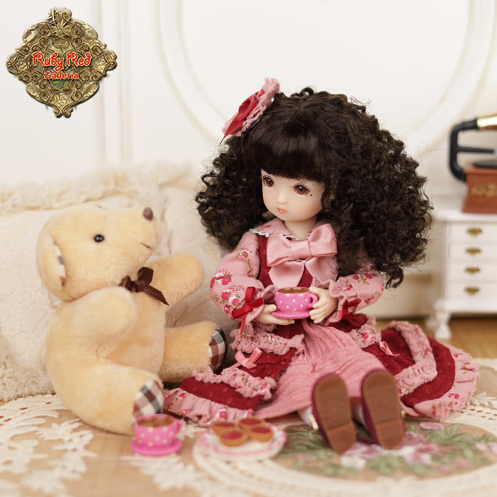 Doll in a pink dress with curly hair sitting next to a teddy bear on a decorative surface.