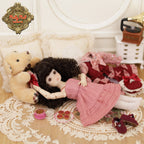 Doll in a pink dress lying on a lace tablecloth with a teddy bear and other toys.