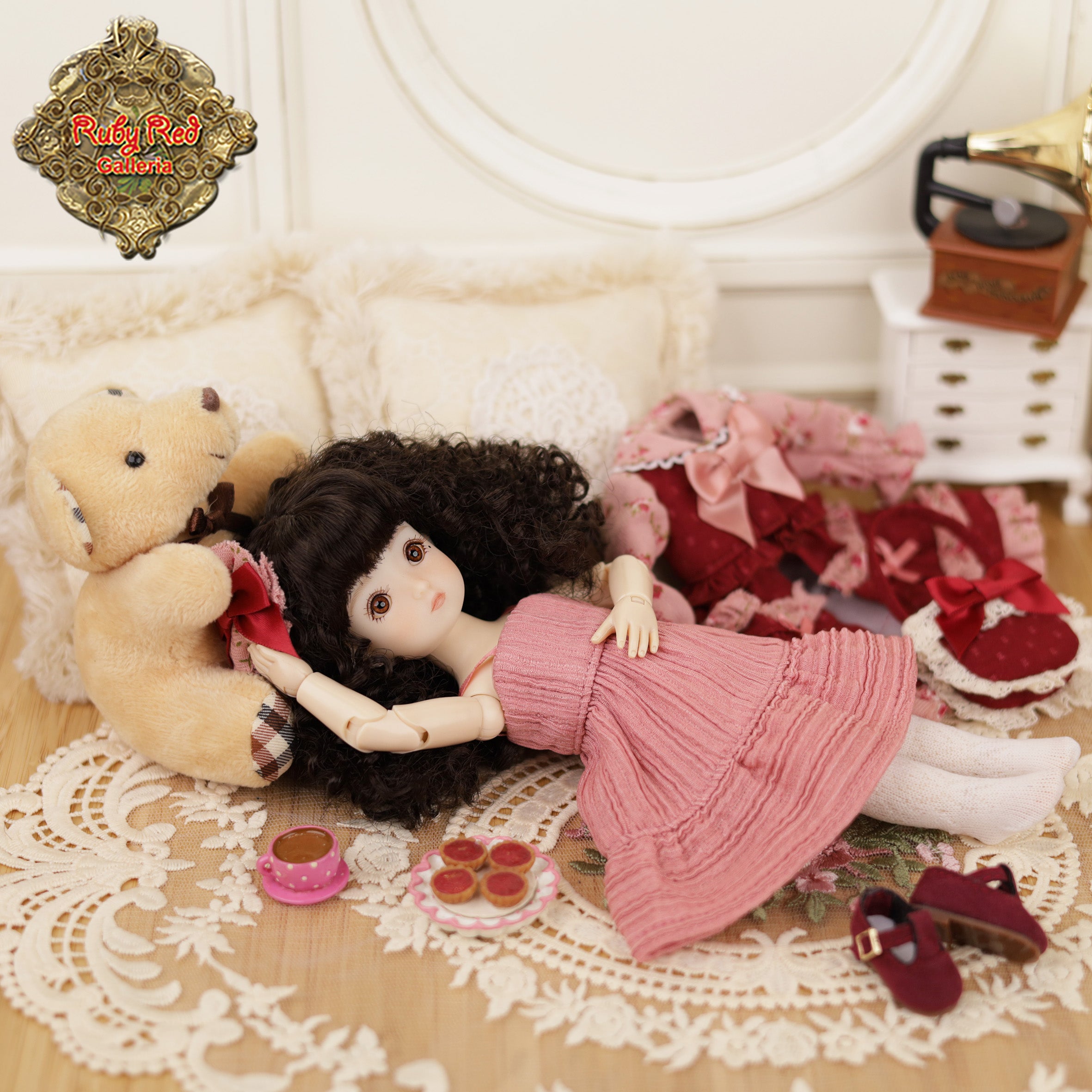 Doll in a pink dress lying on a lace tablecloth with a teddy bear and other toys.
