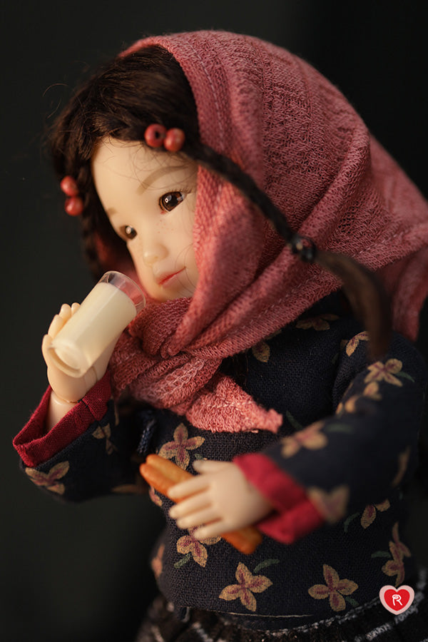Doll wearing a pink scarf and holding a glass of soya milk against a dark background