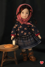 Doll in a dark floral dress with a pink hood standing next to a small wooden table with a glass of milk and fired dough.