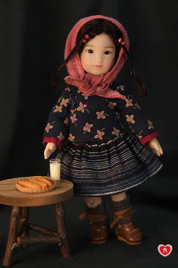 Doll in a dark floral dress with a pink hood standing next to a small wooden table with a glass of milk and fired dough.