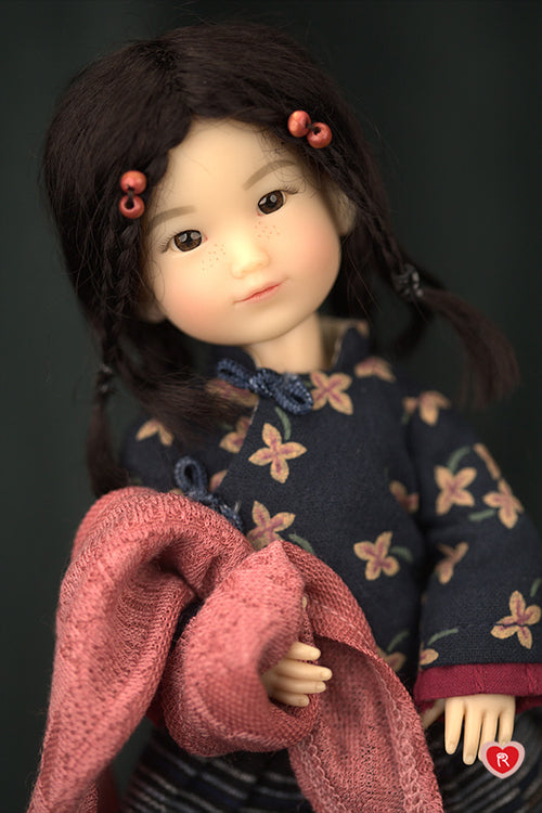 Doll with braided hair and floral dress holding a pink scarf against a dark background