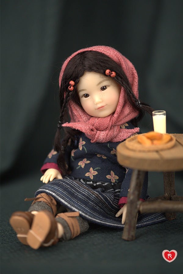 Doll wearing a pink hood and floral dress sitting next to a small table on a dark background