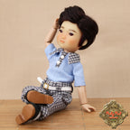 Doll wearing a blue shirt and checkered pants sitting on a wooden surface with a beige background.
