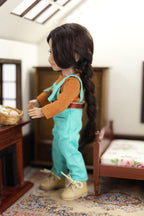 mini Freida  in turquoise overalls and orange shirt standing in a room with furniture and decor.