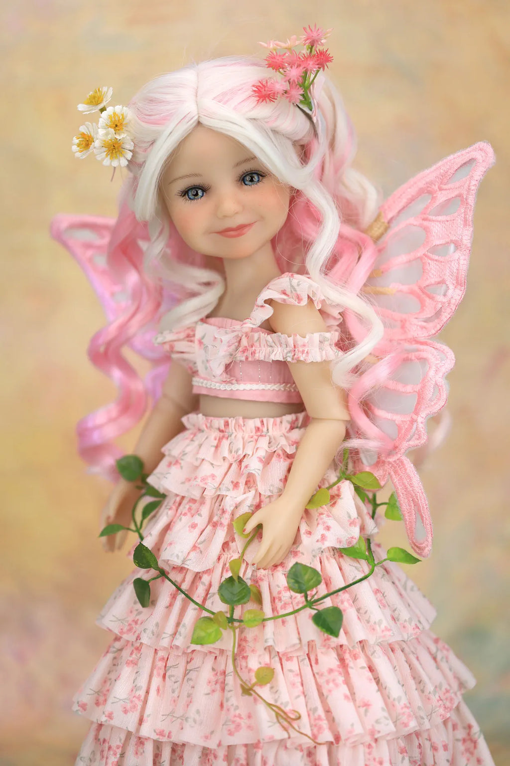 A doll with long white hair, pink fairy wings, and a pink floral dress, posed against a soft beige background with flowers and green leaves around it.