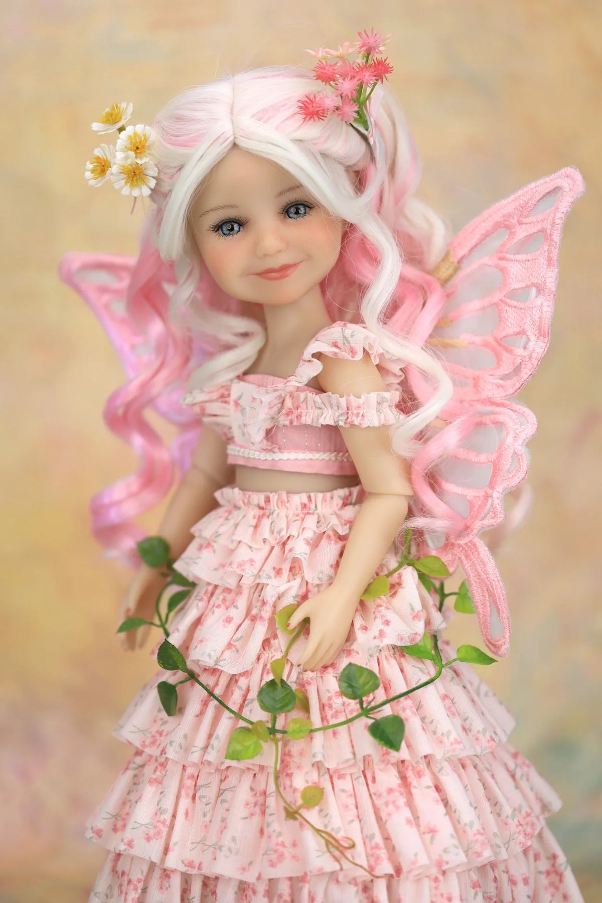 A doll with long white hair, pink fairy wings, and a pink floral dress, posed against a soft beige background with flowers and green leaves around it.