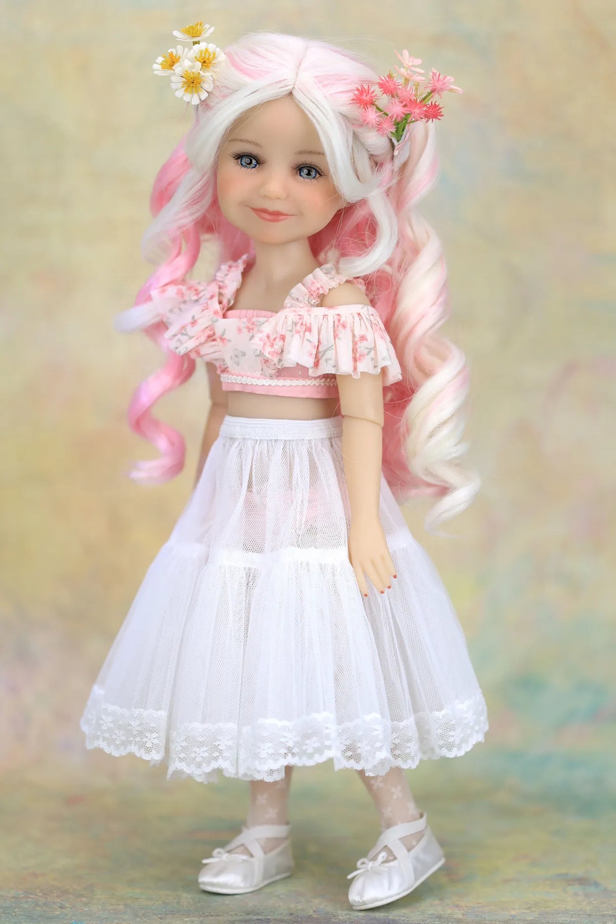 A doll with long wavy white and pink hair, wearing a pink floral off-the-shoulder crop top, a white tiered tulle skirt with lace trim, and white shoes, posed against a soft textured beige background.
