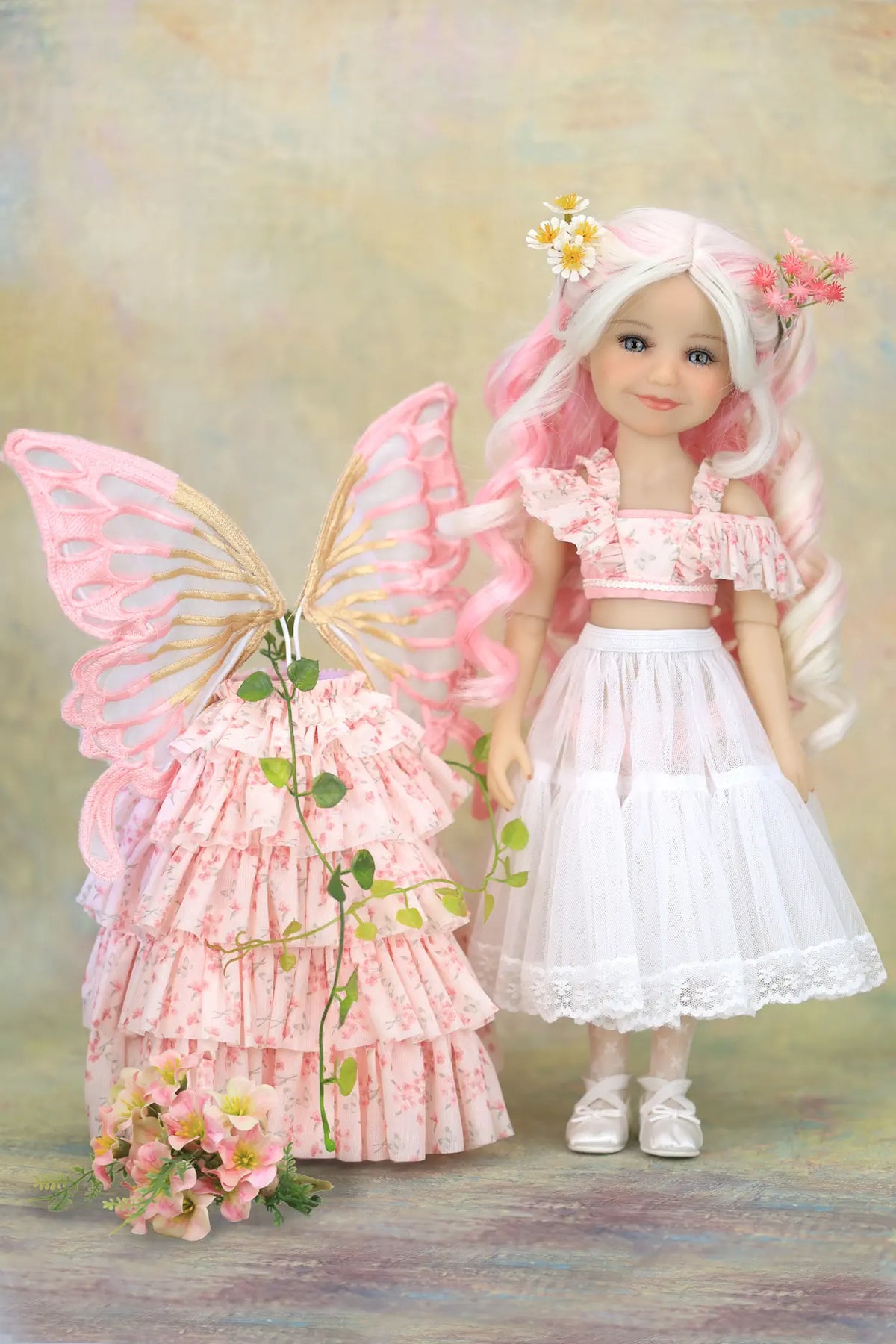 A styled product display featuring a doll with long pink hair and fairy wings next to a pink floral tiered dress with a matching floral headpiece, set against a soft textured beige background.