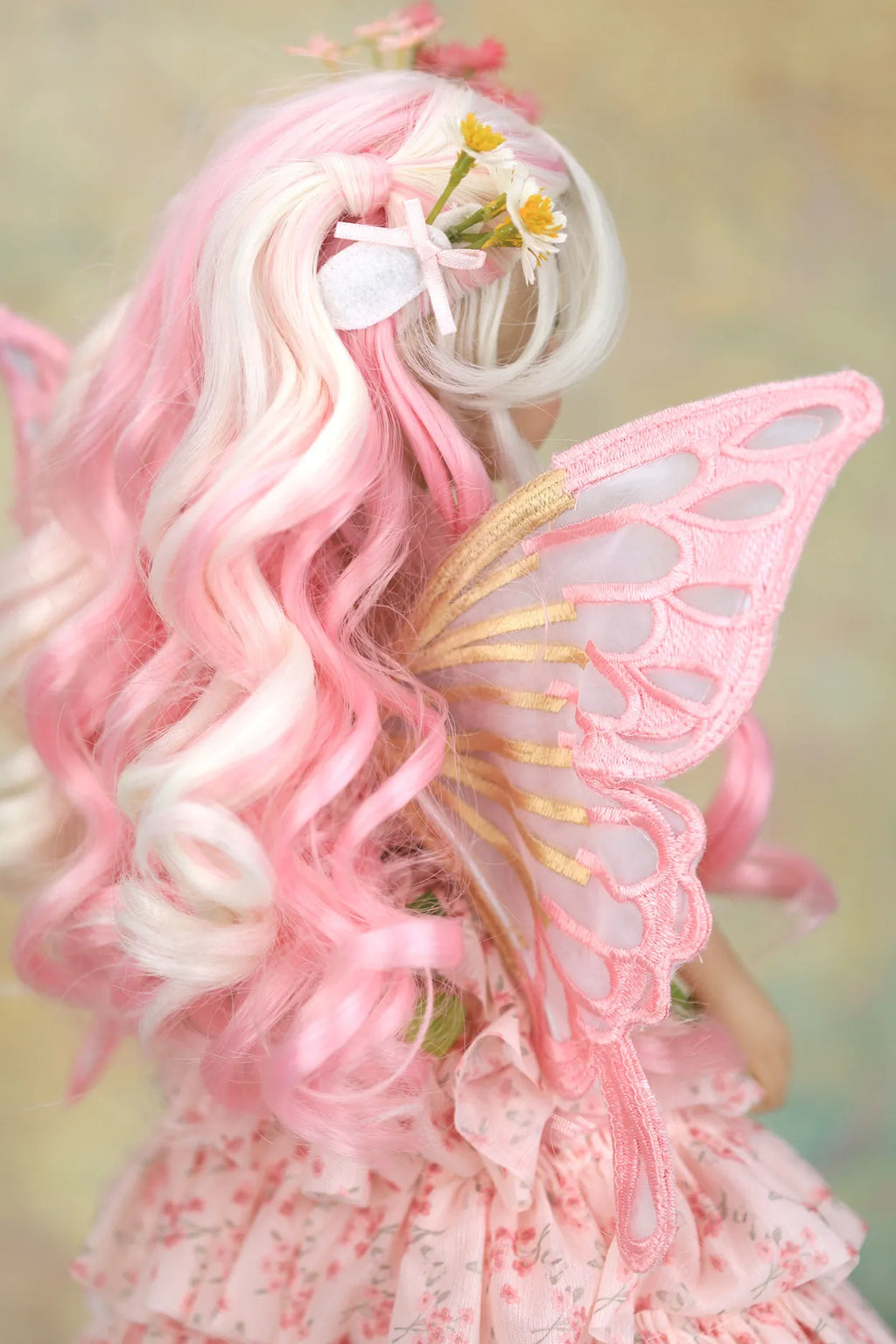 A close-up back view of a doll with long wavy pink and white hair, small yellow daisy flowers in the hair, pink lace wings with gold trim, and a pink floral dress against a soft blurred background.