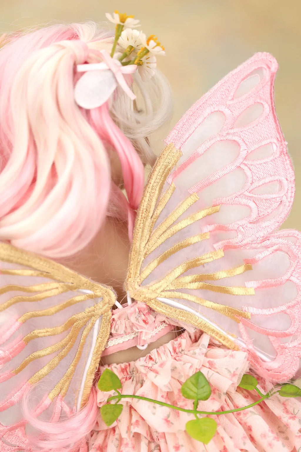 A close-up of a pink fairy costume accessory set, featuring sheer pink wings with gold trim, a pink ruffled dress, and pink hair with small white flowers.