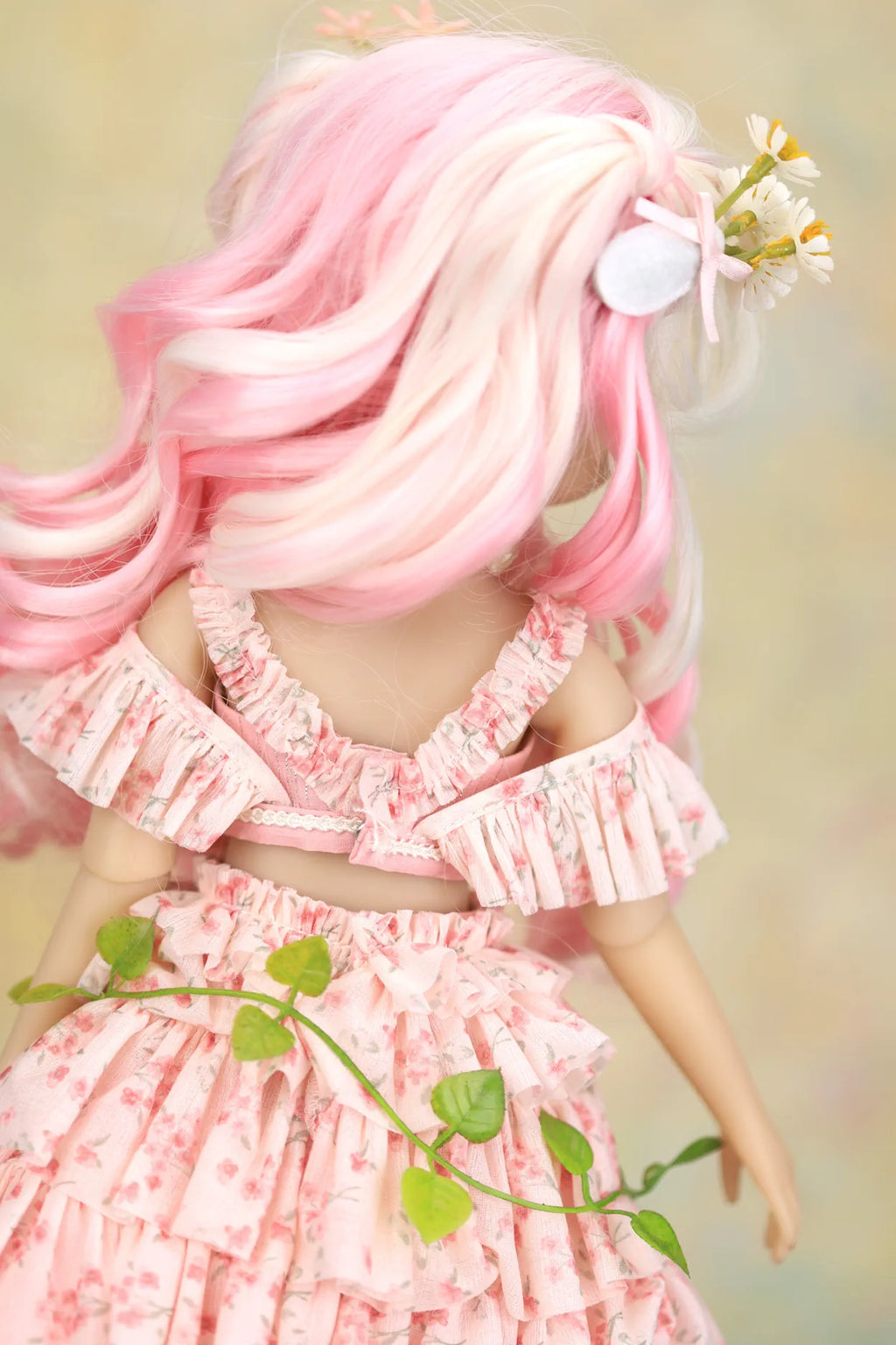 A doll viewed from behind wearing a pink floral off-the-shoulder ruffled dress, with long pink and white wavy hair and a small white flower accessory in its hair, set against a soft beige background.