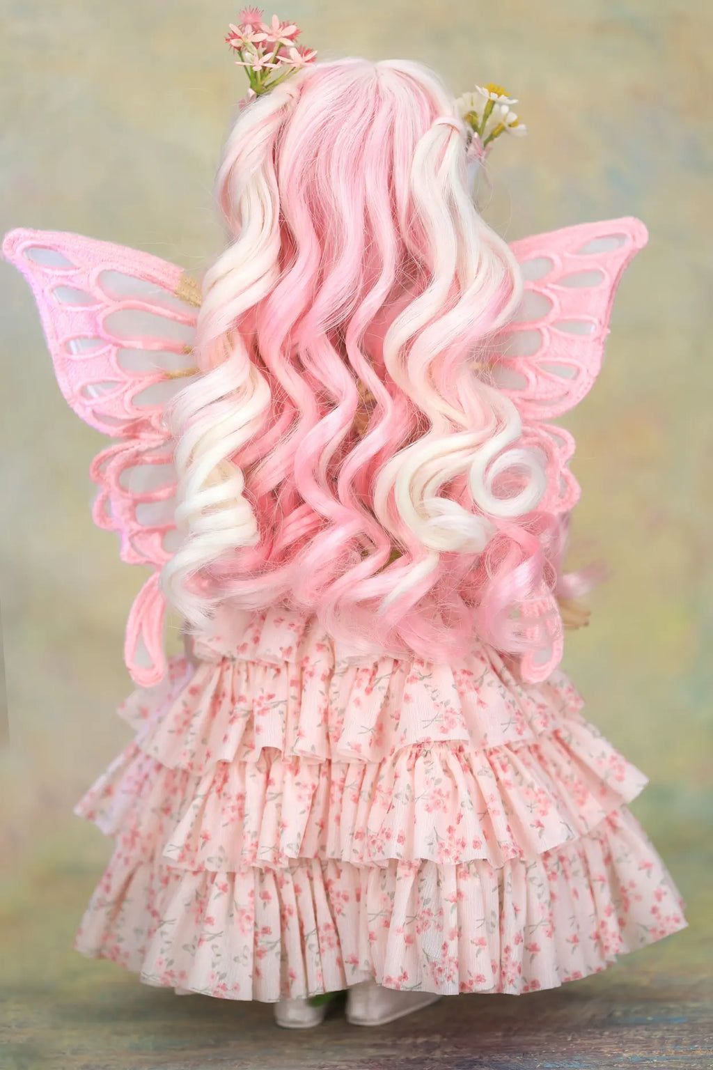 A doll viewed from the back wearing a pink fairy costume, featuring long wavy pink and white hair, pink wings, and a ruffled pink floral dress, set against a soft textured background.