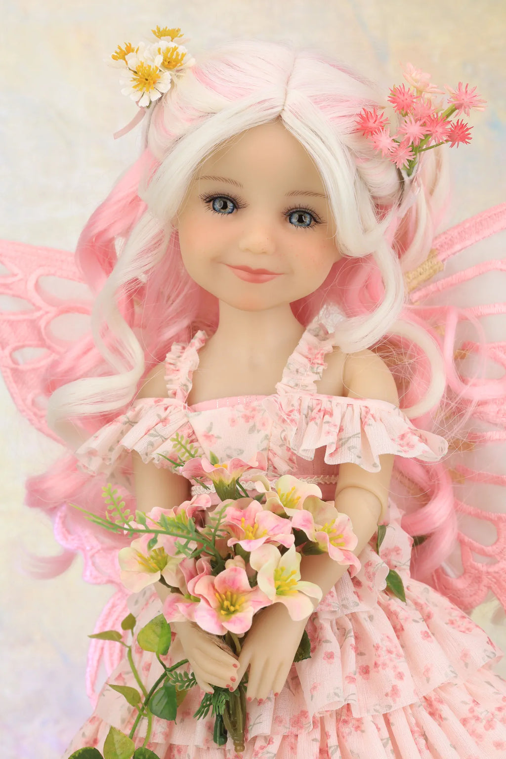 A pink fairy doll with long white hair, delicate wings, and a floral dress, holding a bouquet of pink and yellow flowers against a soft beige background.