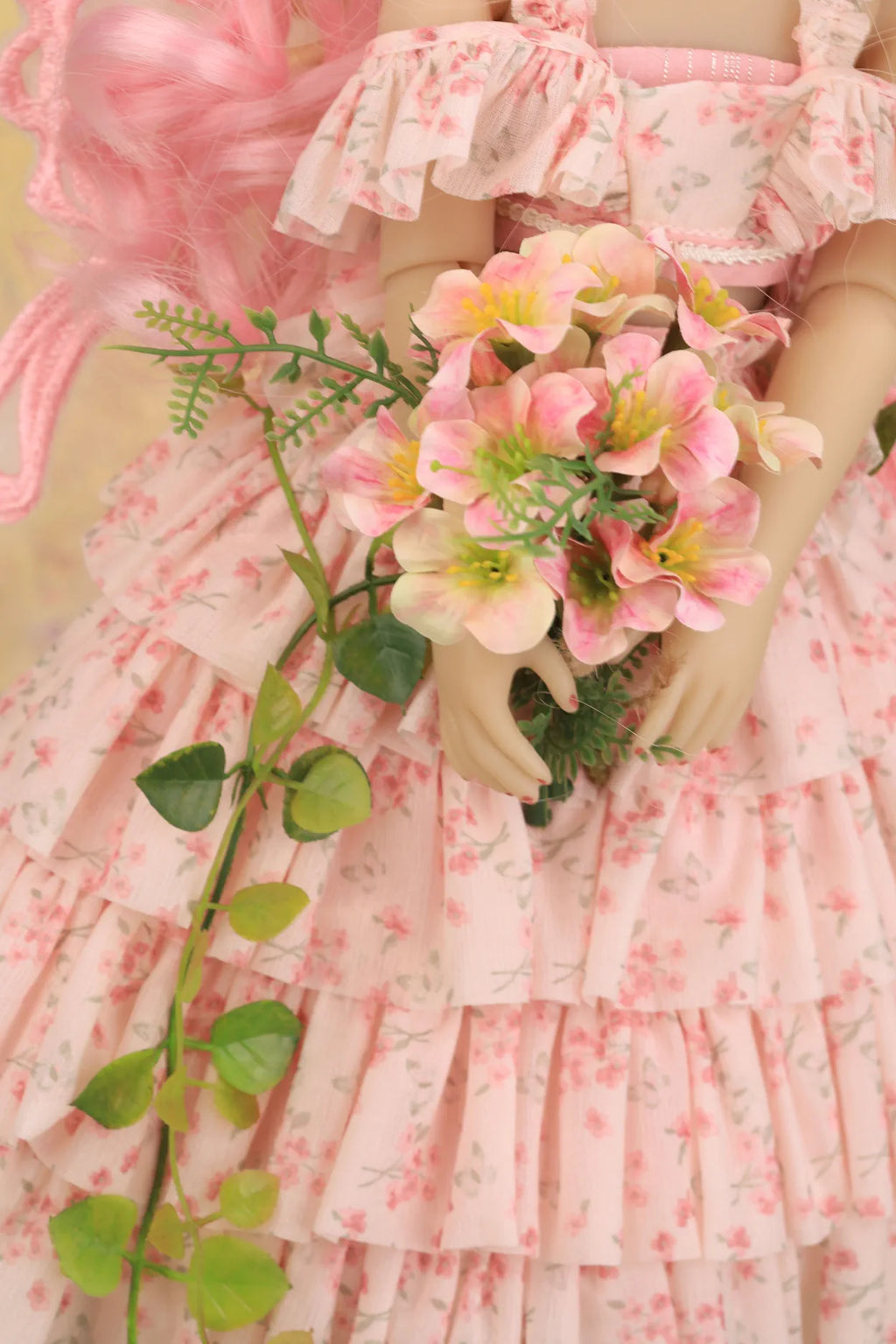 A close-up of a doll wearing a pink floral off-the-shoulder ruffled dress and holding a bouquet of pink flowers with green leaves.