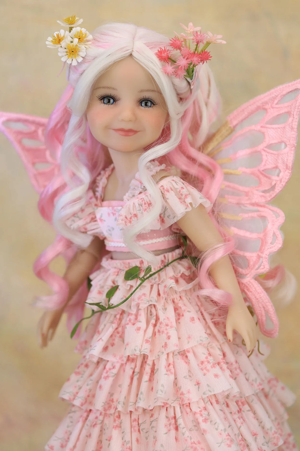 A doll with long wavy white and pink hair, wearing a pink floral fairy dress and translucent pink wings, posed against a soft beige background.