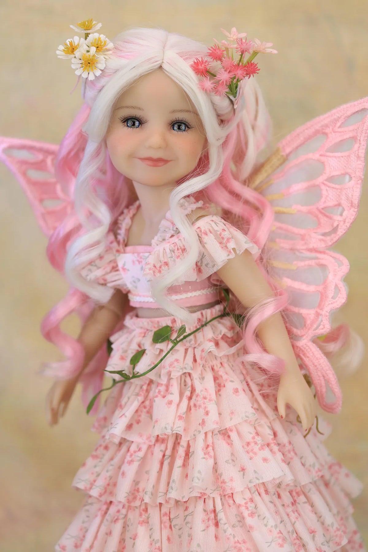 A doll with long wavy white and pink hair, wearing a pink floral fairy dress and translucent pink wings, posed against a soft beige background.