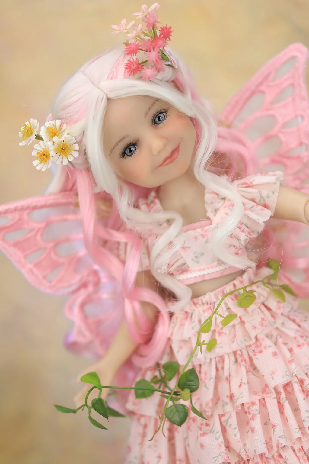 A close-up of a doll or figurine with long white hair, pink fairy wings, and a pink floral dress, styled with small flowers in her hair and a green vine in her hand against a soft beige background.
