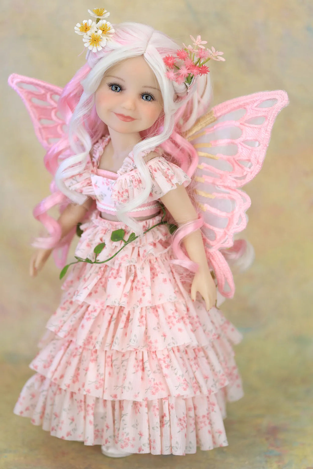 A fairy doll with long white hair, pink wings, and a pink floral dress, posed against a soft beige background.