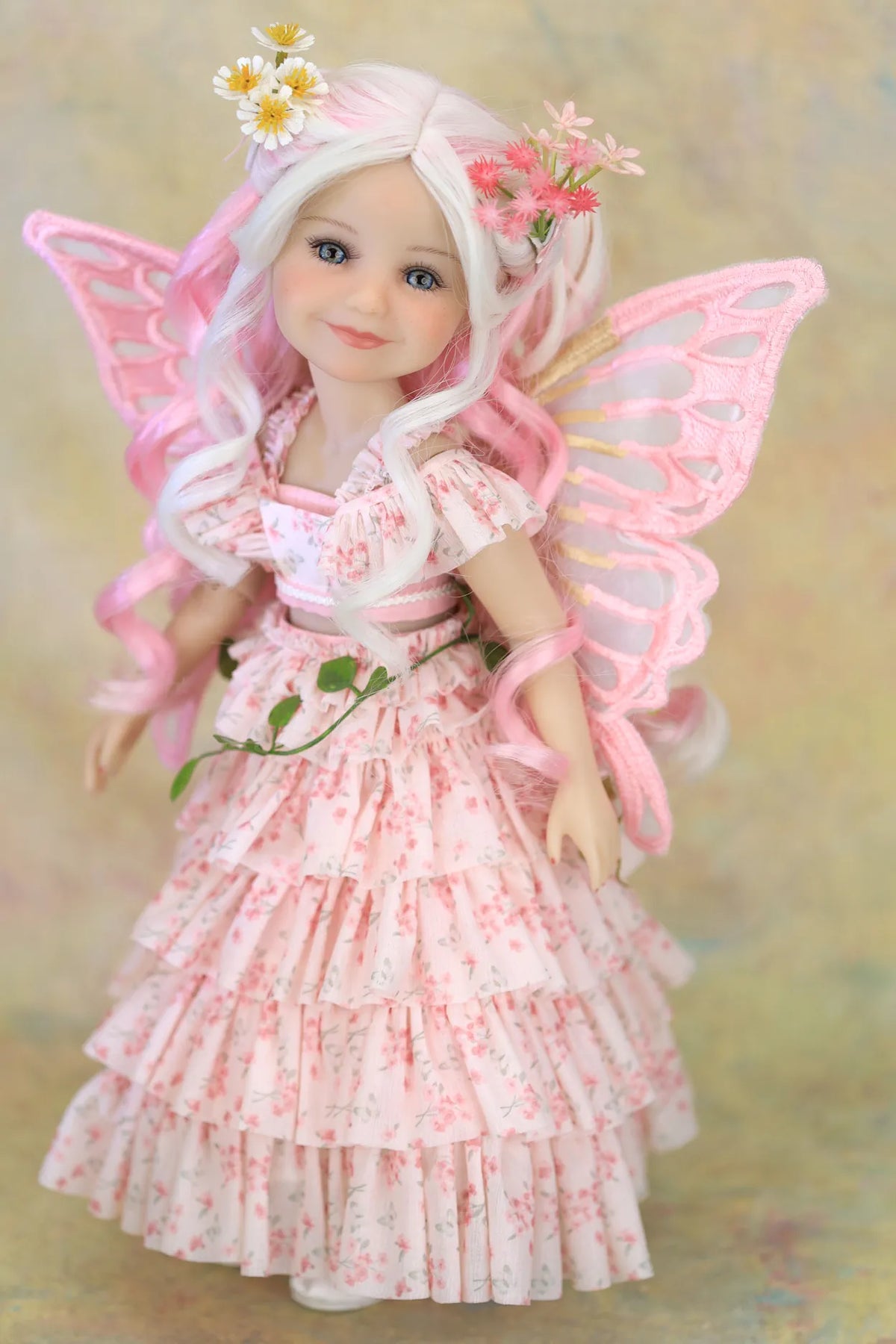 A fairy doll with long white hair, pink wings, and a pink floral dress, posed against a soft beige background.