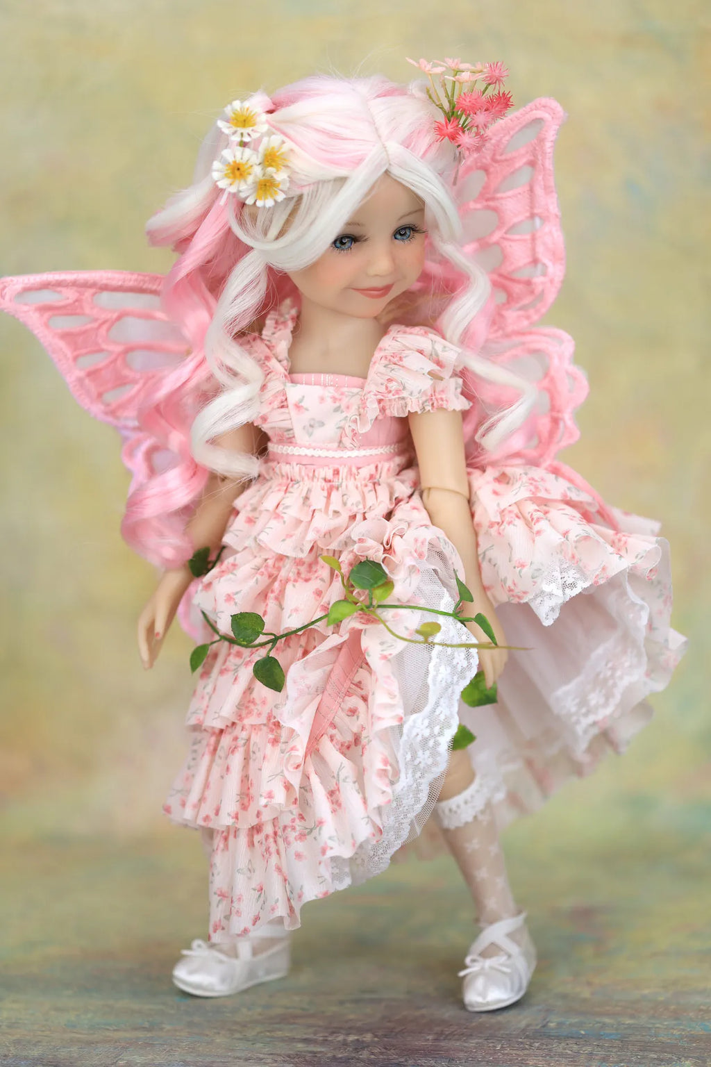 A doll or figurine dressed as a fairy, with long white hair, pink wings, and a pink floral dress, posed against a soft pastel background.