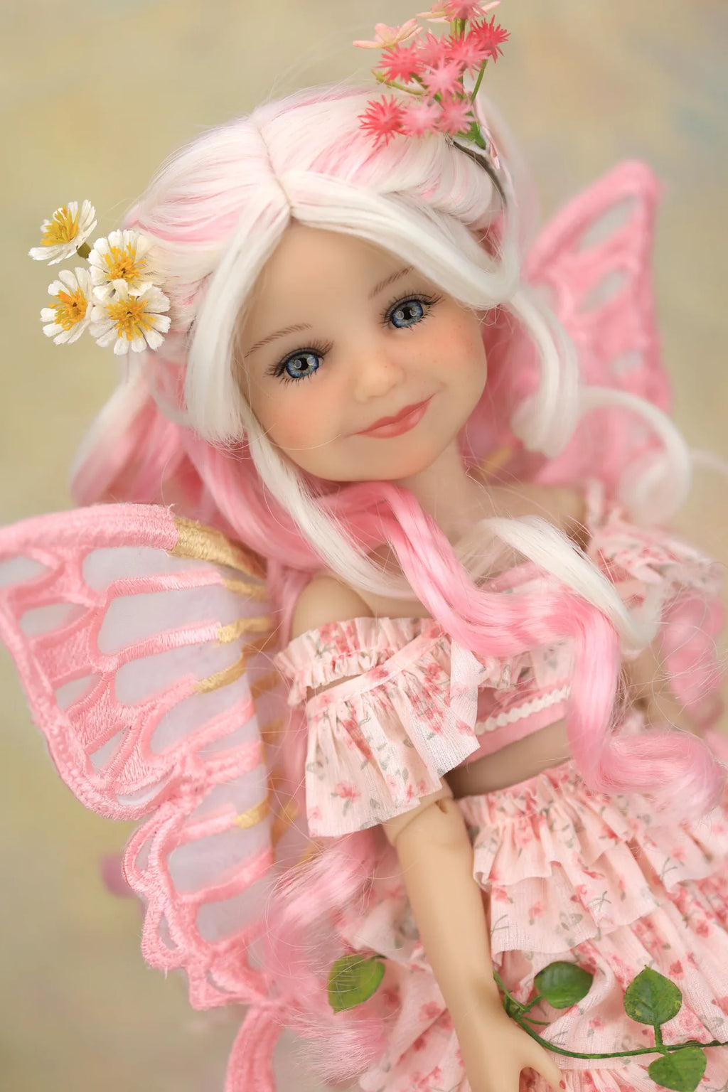 A close-up of a doll with long white and pink hair, wearing a pink fairy-style dress and translucent pink wings, with small flower and leaf decorations, against a soft beige background.