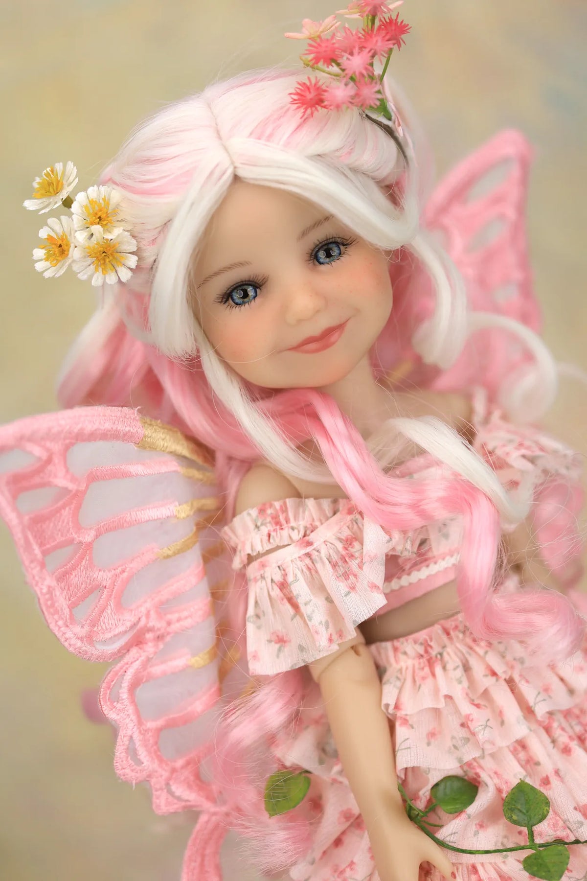 A close-up of a doll with long white and pink hair, wearing a pink fairy-style dress and translucent pink wings, with small flower and leaf decorations, against a soft beige background.