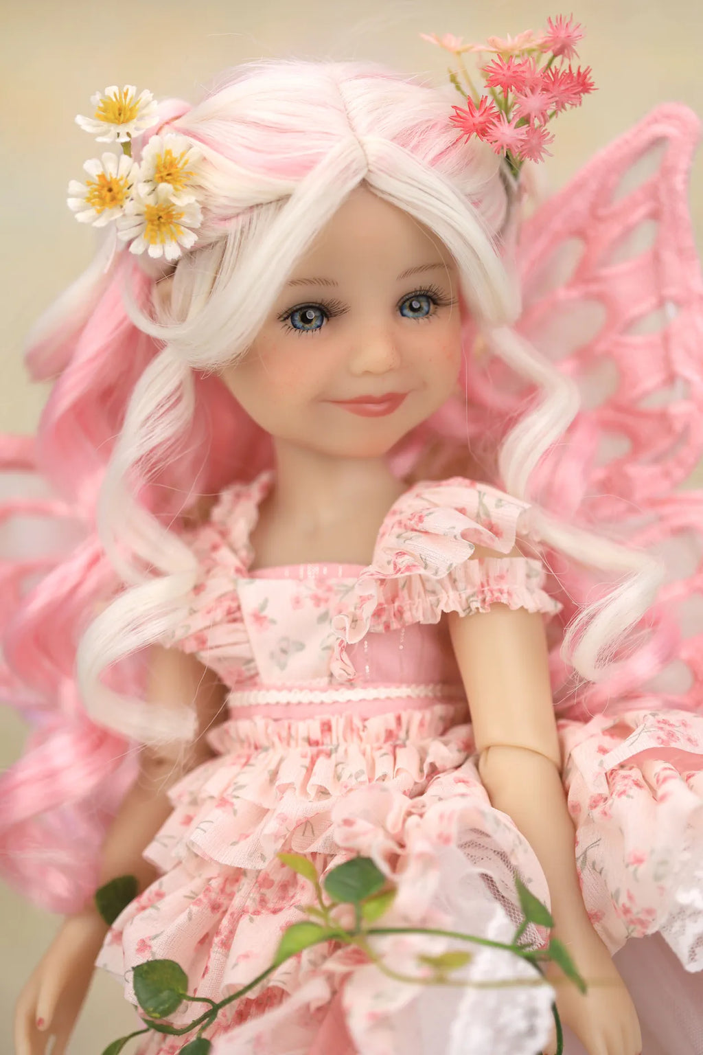 A doll with long white hair, pink fairy wings, and a pink floral dress, styled with daisy and pink flowers in her hair, set against a soft beige background.