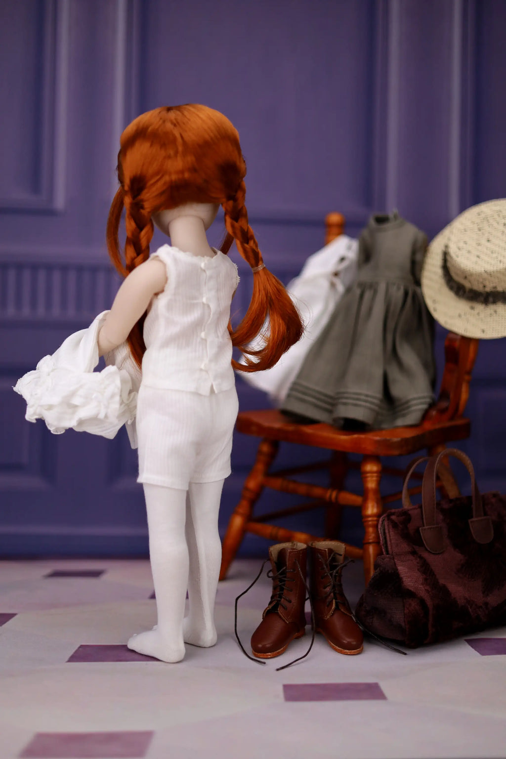 Doll with red hair in pigtails wearing a white dress, standing next to a chair with a green dress and a straw hat on a purple background.