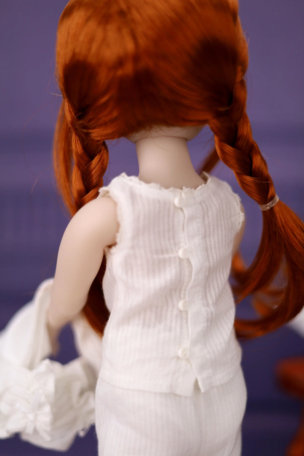 Doll with red hair in pigtails wearing a white dress against a purple background