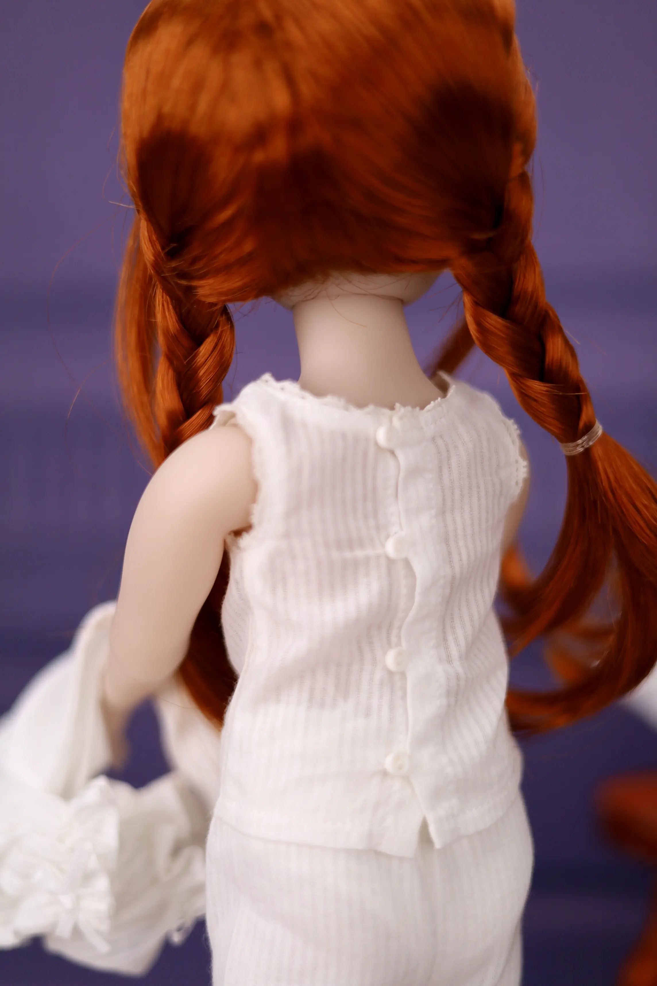 Doll with red hair in pigtails wearing a white dress against a purple background