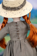 Doll with braided red hair wearing a straw hat and gray dress against a blurred natural background