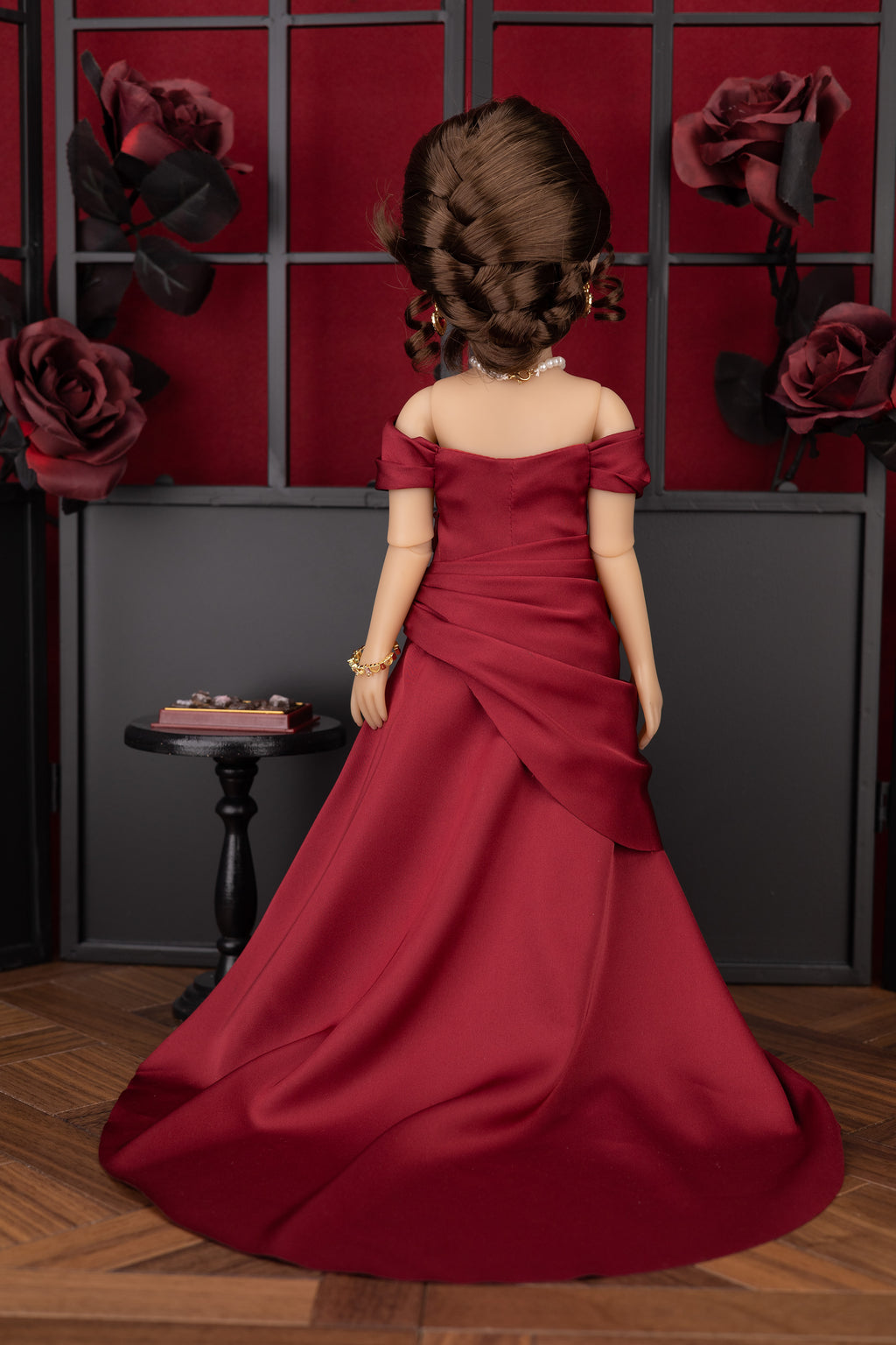 Doll in a red dress standing in front of a decorative wall with roses.