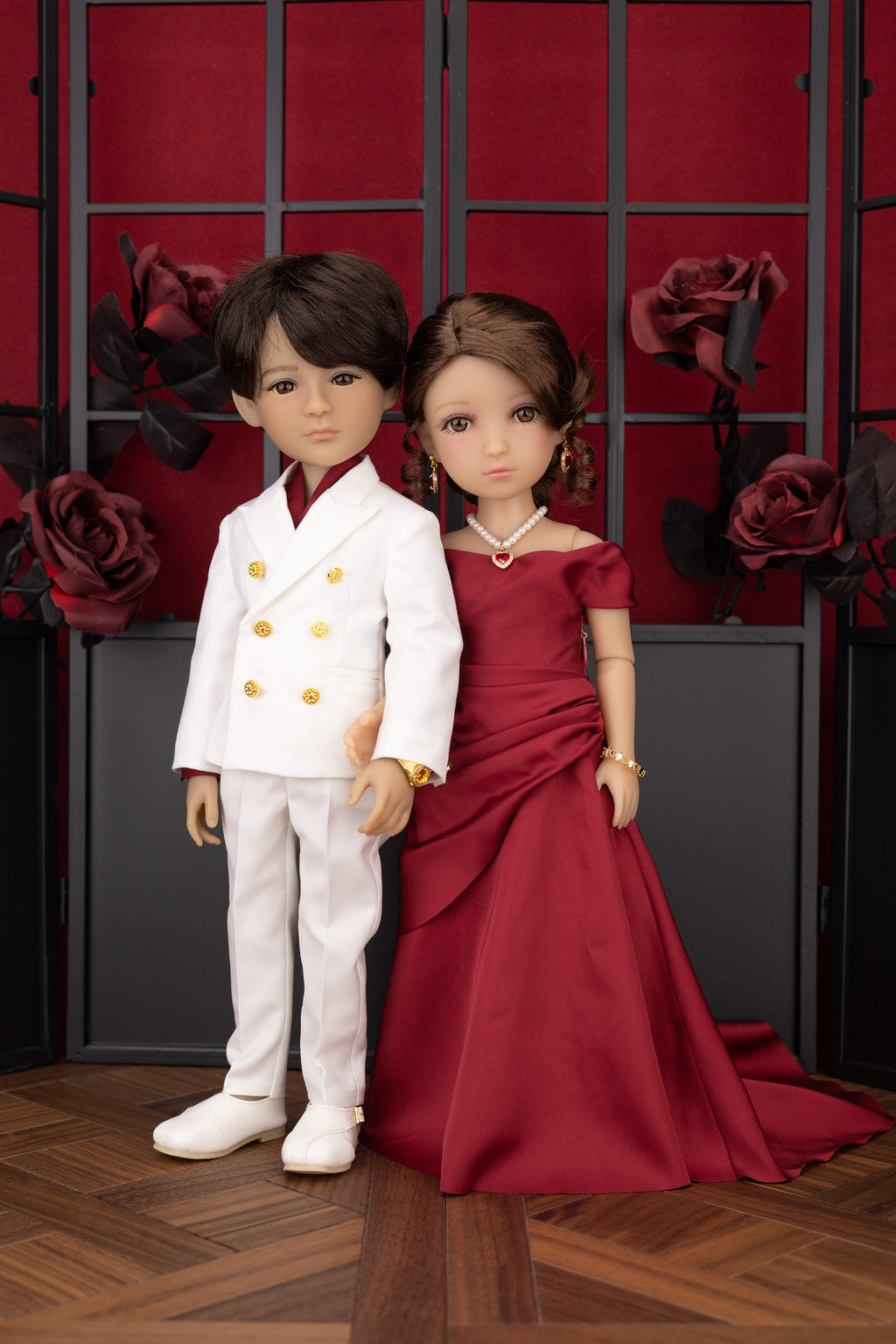 Two dolls, one in a white outfit with gold buttons and the other in a red dress, standing against a decorative background with red roses.