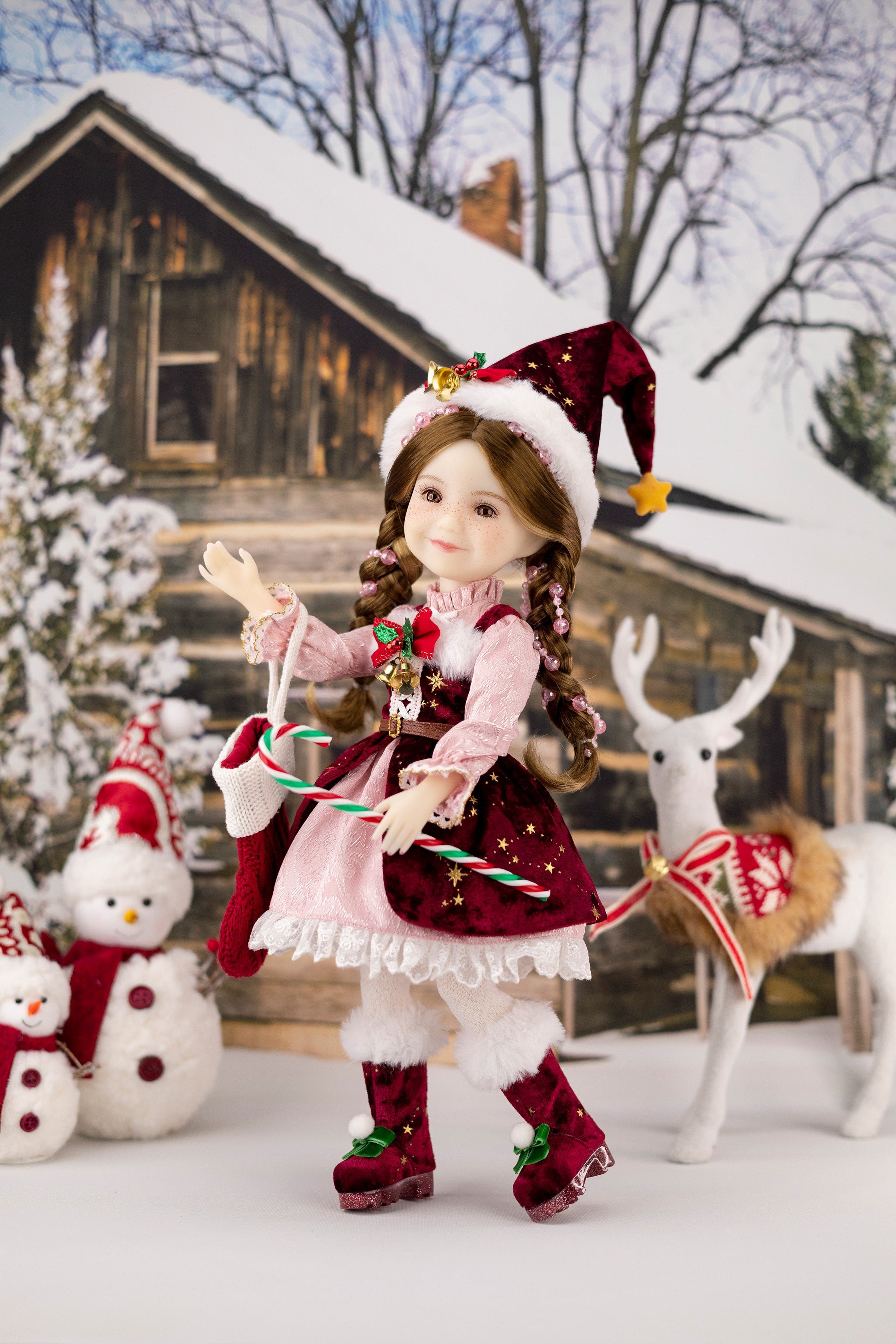 Christmas-themed doll with a snowy background featuring a cabin and deer.