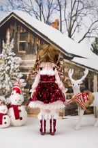 Doll in a festive outfit standing in front of a snowy cabin with Christmas decorations.