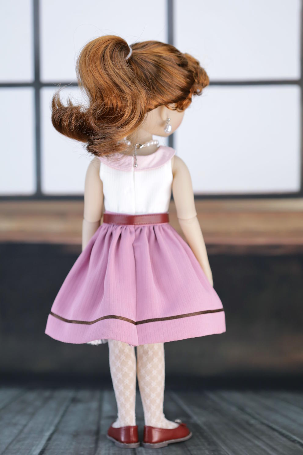 Doll wearing a pink dress with a red belt on a wooden floor.