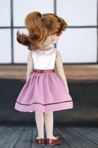 Doll wearing a pink dress with a red belt on a wooden floor.