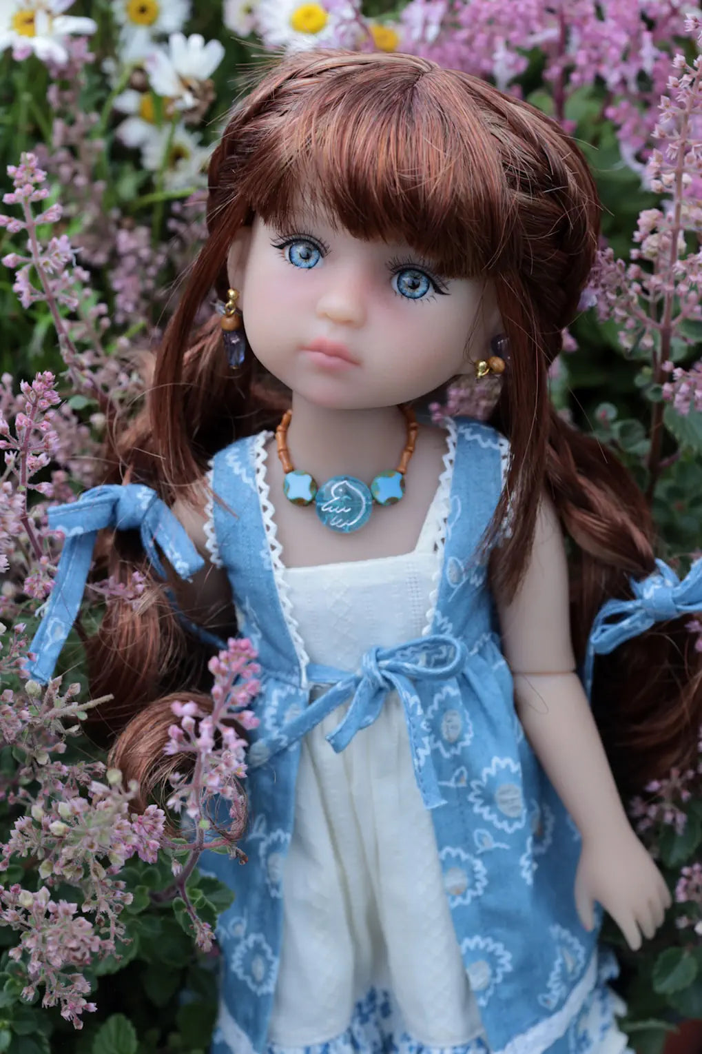Doll with long brown hair and blue eyes, wearing a blue dress and white top, surrounded by flowers.