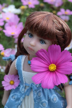  Doll with brown hair and blue eyes holding a pink flower in front of its face, surrounded by flowers.