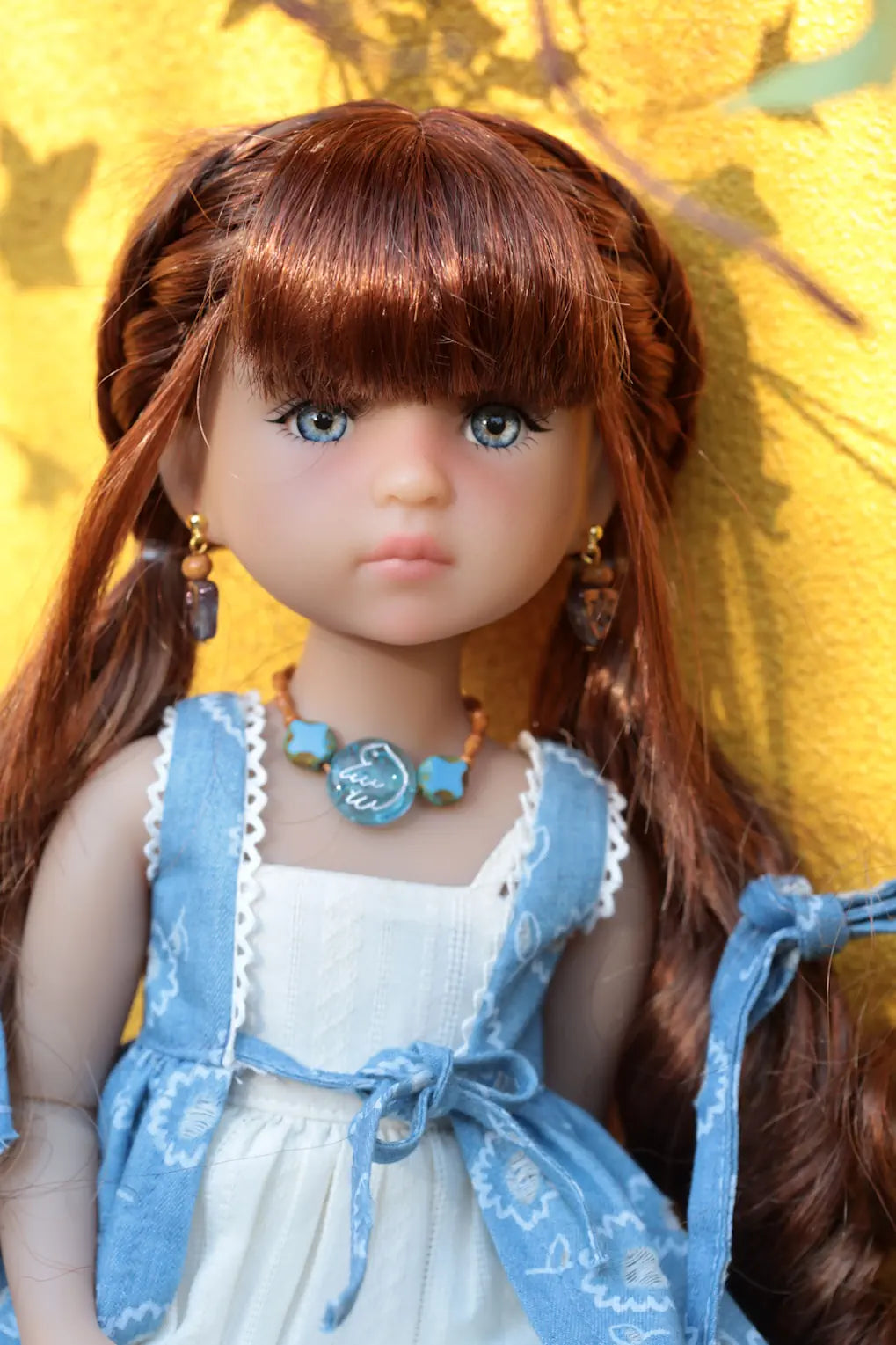 Doll with brown hair styled in pigtails, wearing a white dress and blue jacket against a yellow background.