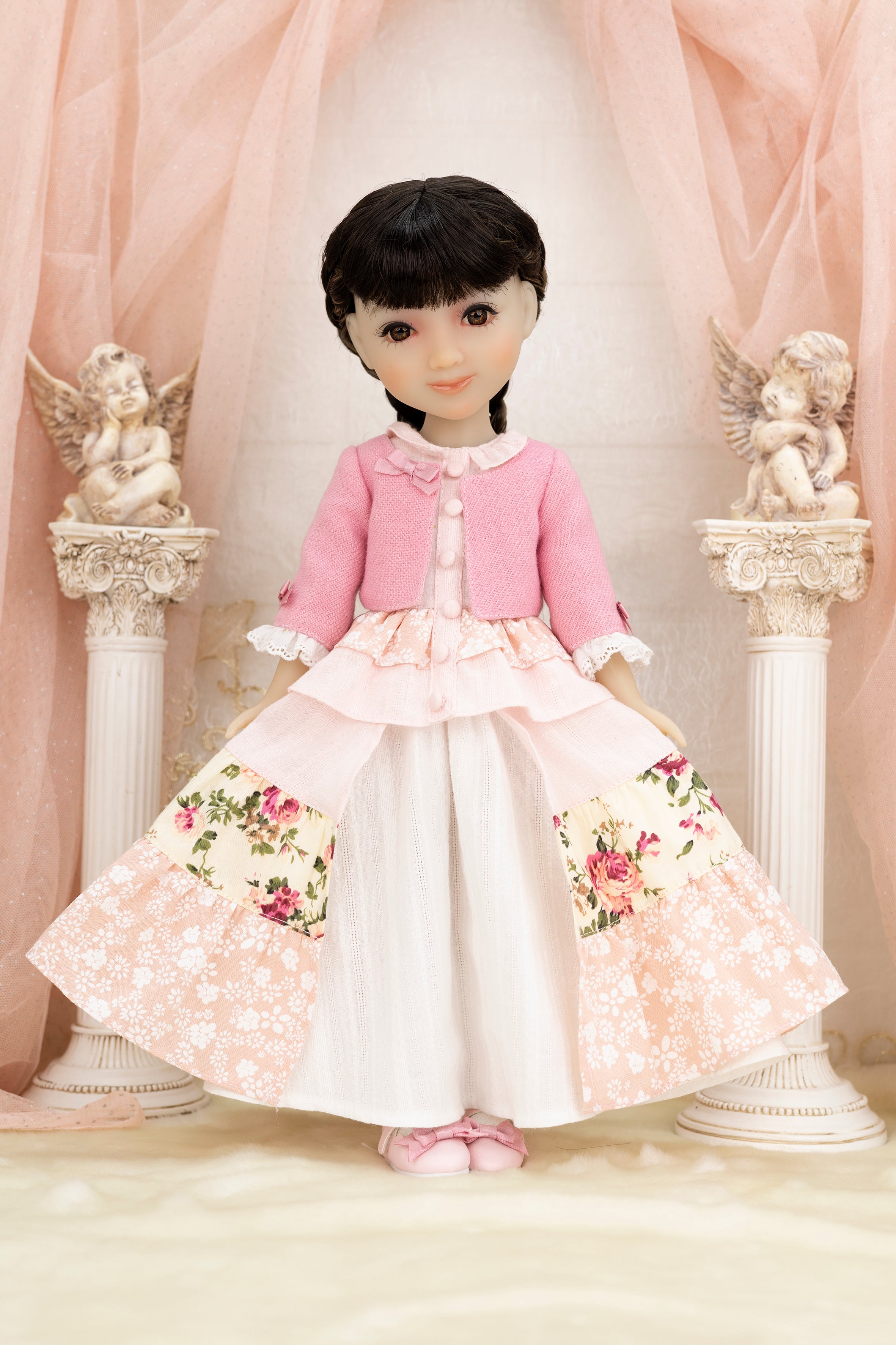 Doll wearing a pink cardigan and floral dress with cherub statues in the background