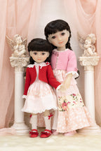 Two dolls in formal attire standing in front of decorative columns with angel statues.