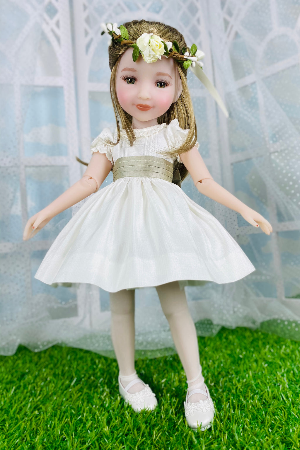 Doll in a white dress with a floral headband standing on grass with a white archway in the background