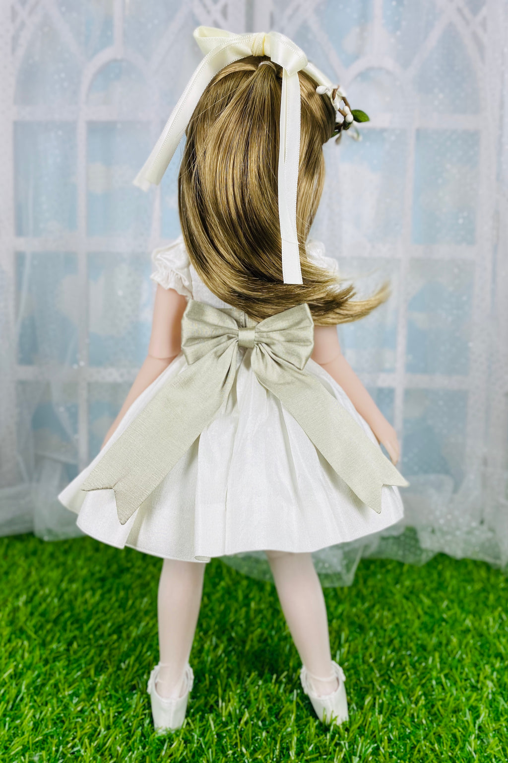 Doll in a white dress with a large bow, standing on grass with a light blue curtain background