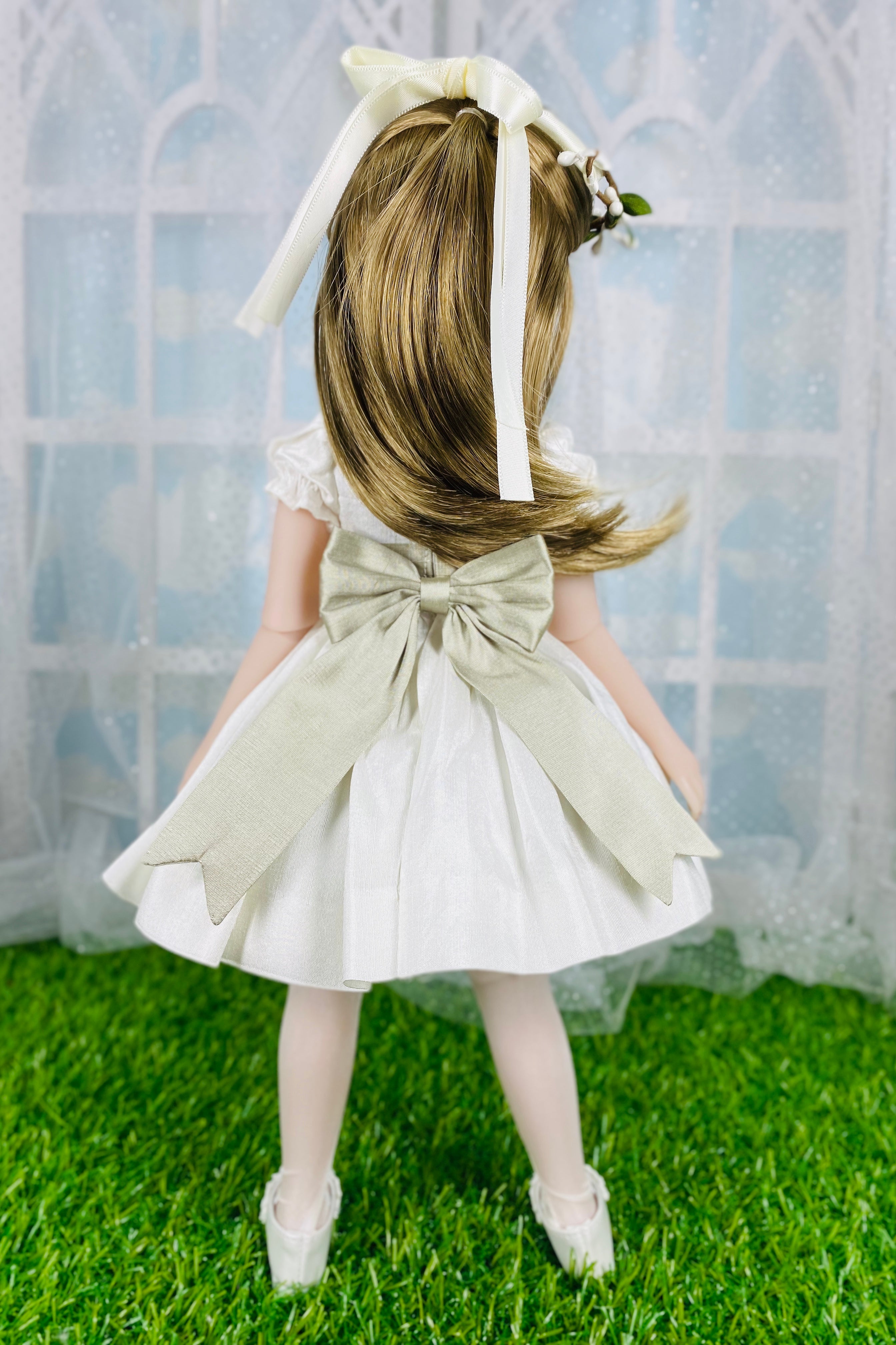 Doll in a white dress with a large bow, standing on grass with a light blue curtain background