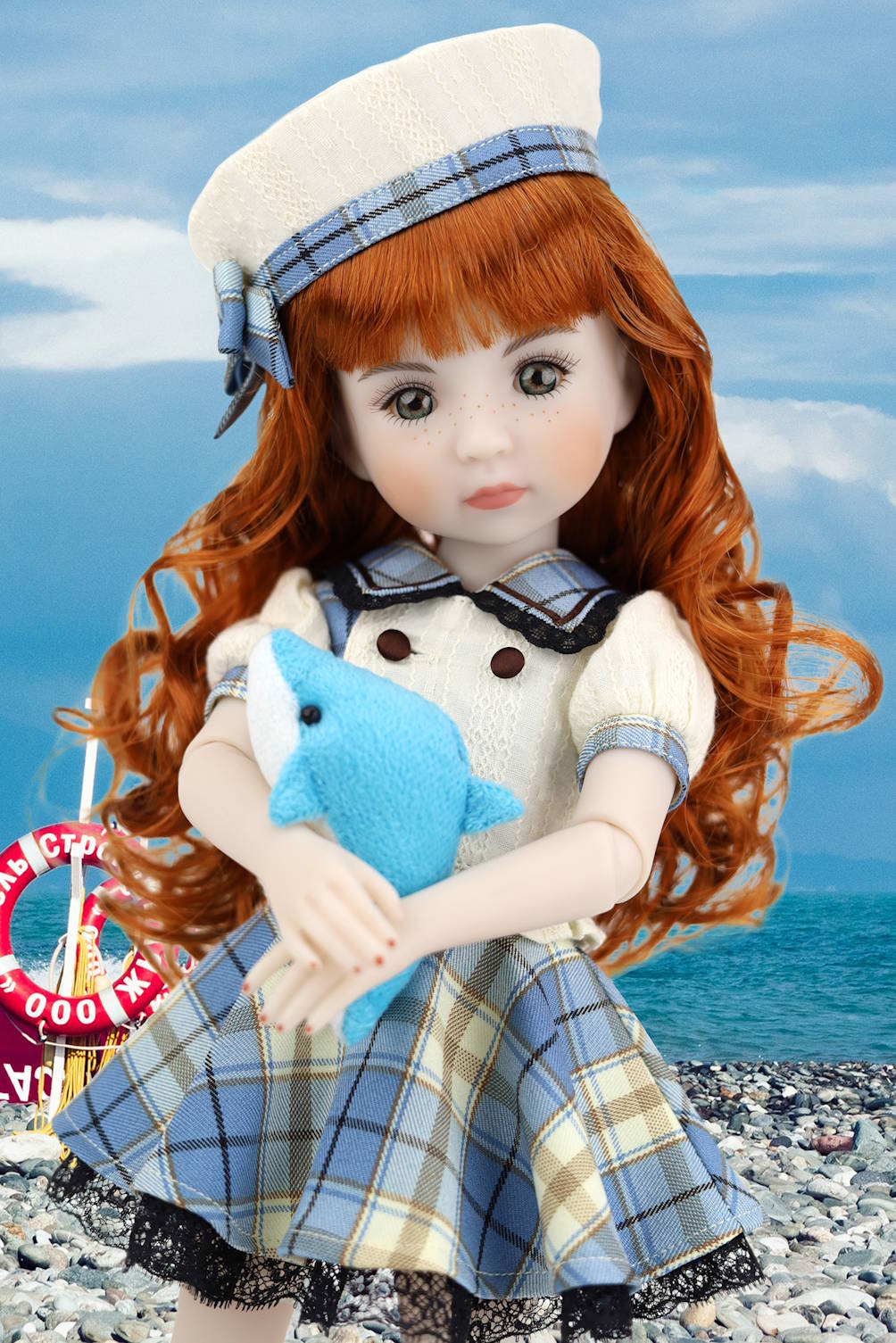Doll with red hair wearing a sailor outfit holding a blue plush toy on a beach.