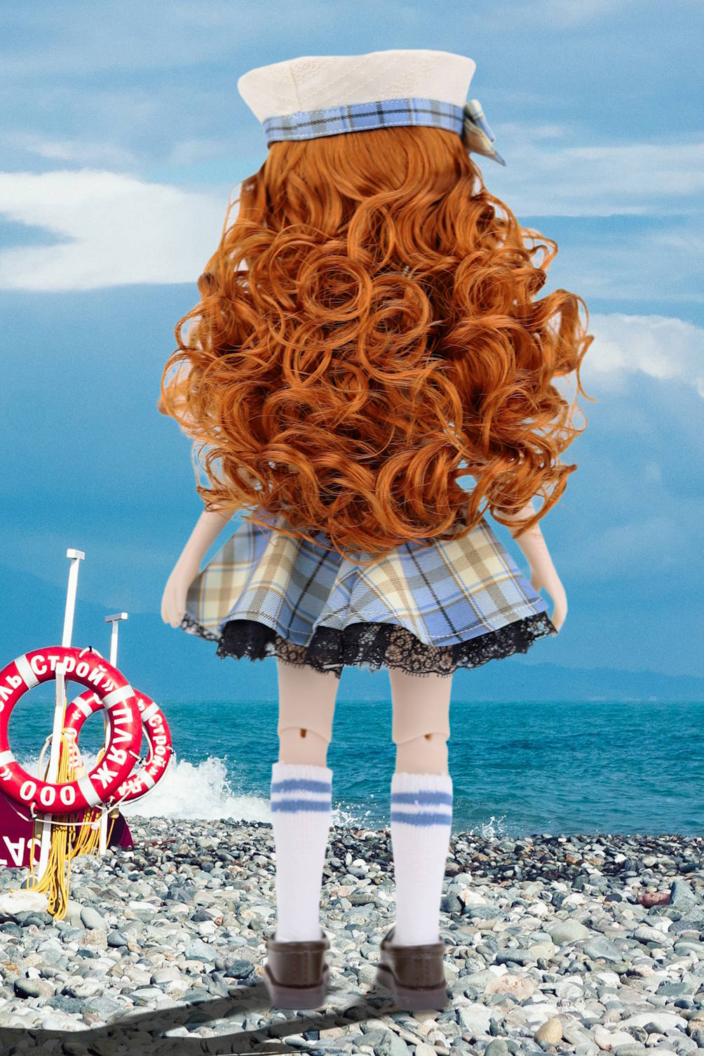 Doll in a sailor outfit with red hair standing on a rocky beach with ocean and sky in the background.